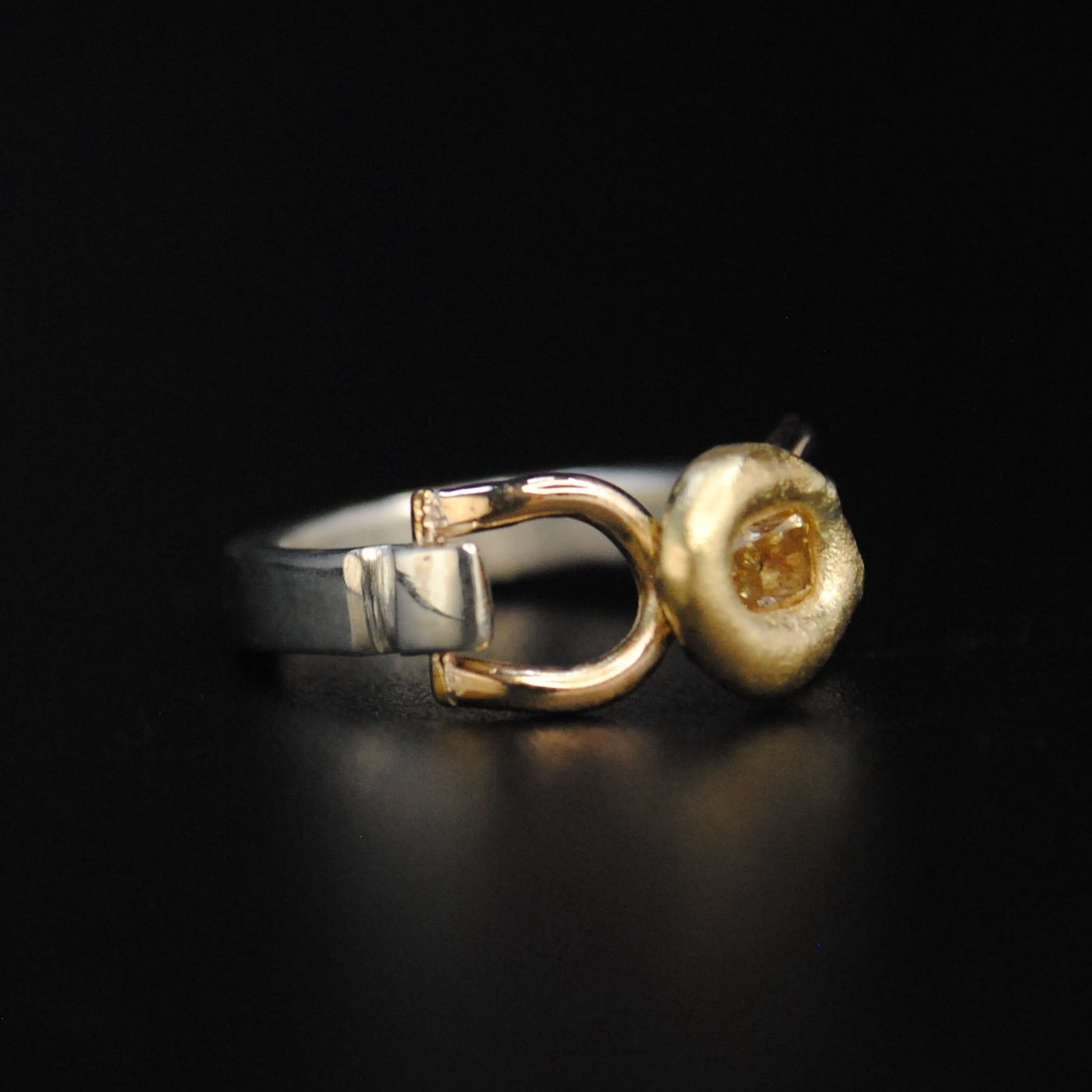 A two-tone metal ring with a yellow gold flower-shaped setting and a silver-toned band, displayed on a reflective black surface.
