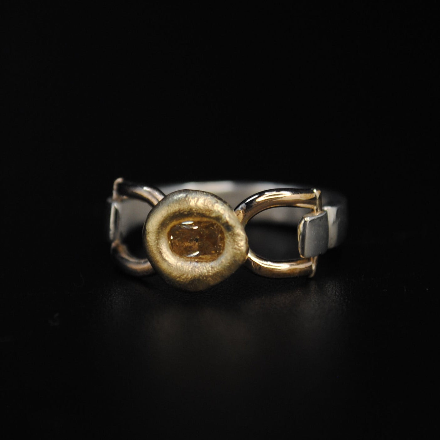 A two-tone metal ring with a textured gold center and silver-toned bands, displayed on a solid black reflective surface.