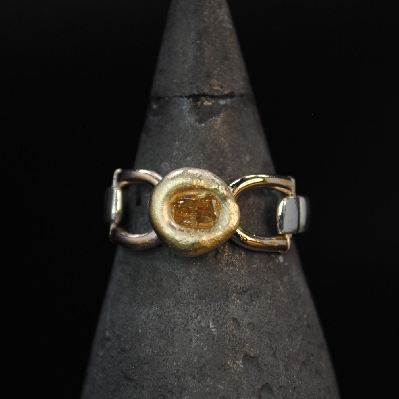 A gold ring with a textured circular center and small white inlay details, displayed on a dark metal cone stand against a black background.