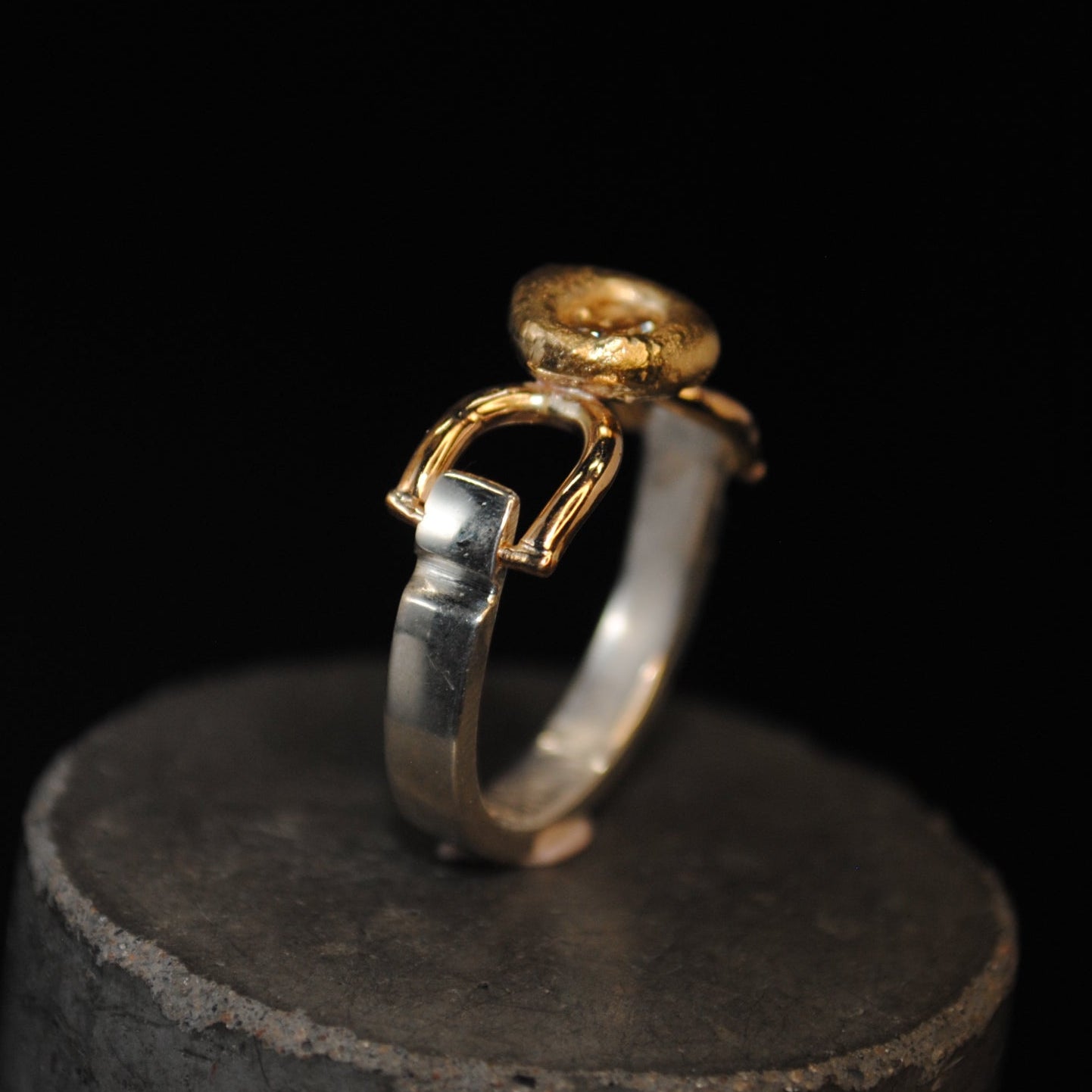 A two-tone ring with a gold-colored top and band details and a silver-colored shank, displayed on a dark cylindrical stand against a solid black background.