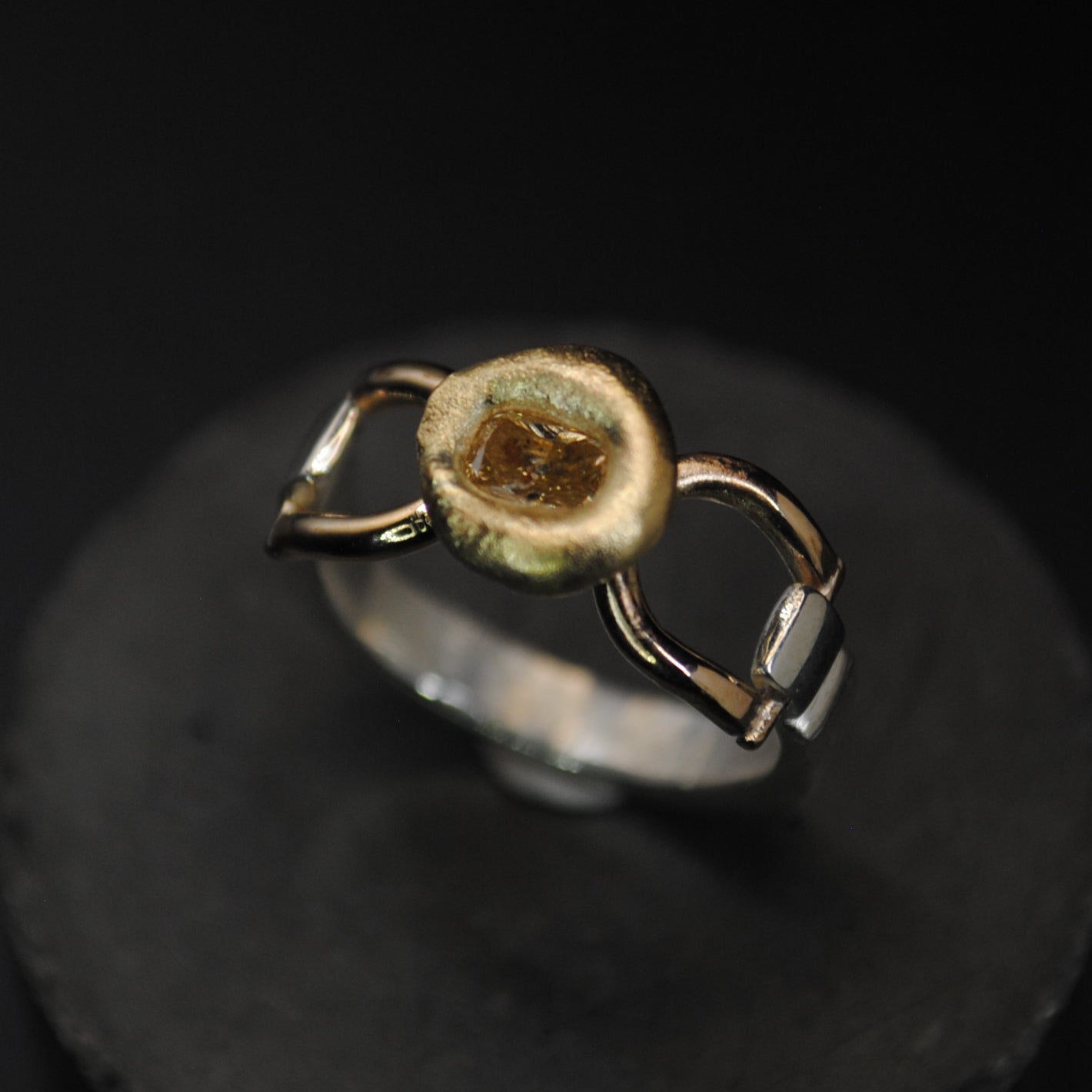 A silver ring with a gold-toned circular center and small gold side accents, displayed on a round black stand against a black background.