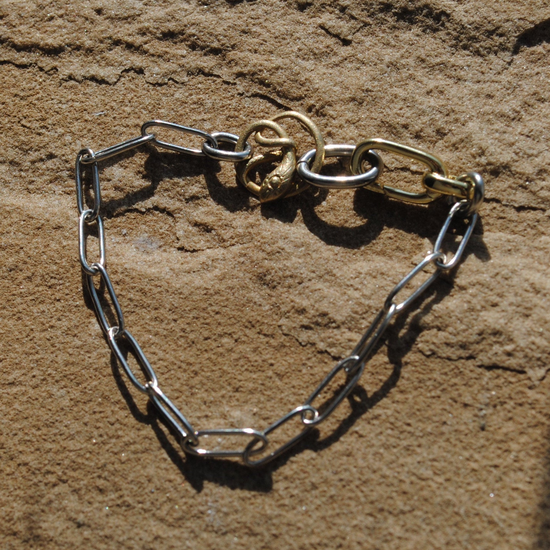 A silver chain link bracelet with a gold-toned lobster clasp, displayed on a textured tan stone surface under direct sunlight.