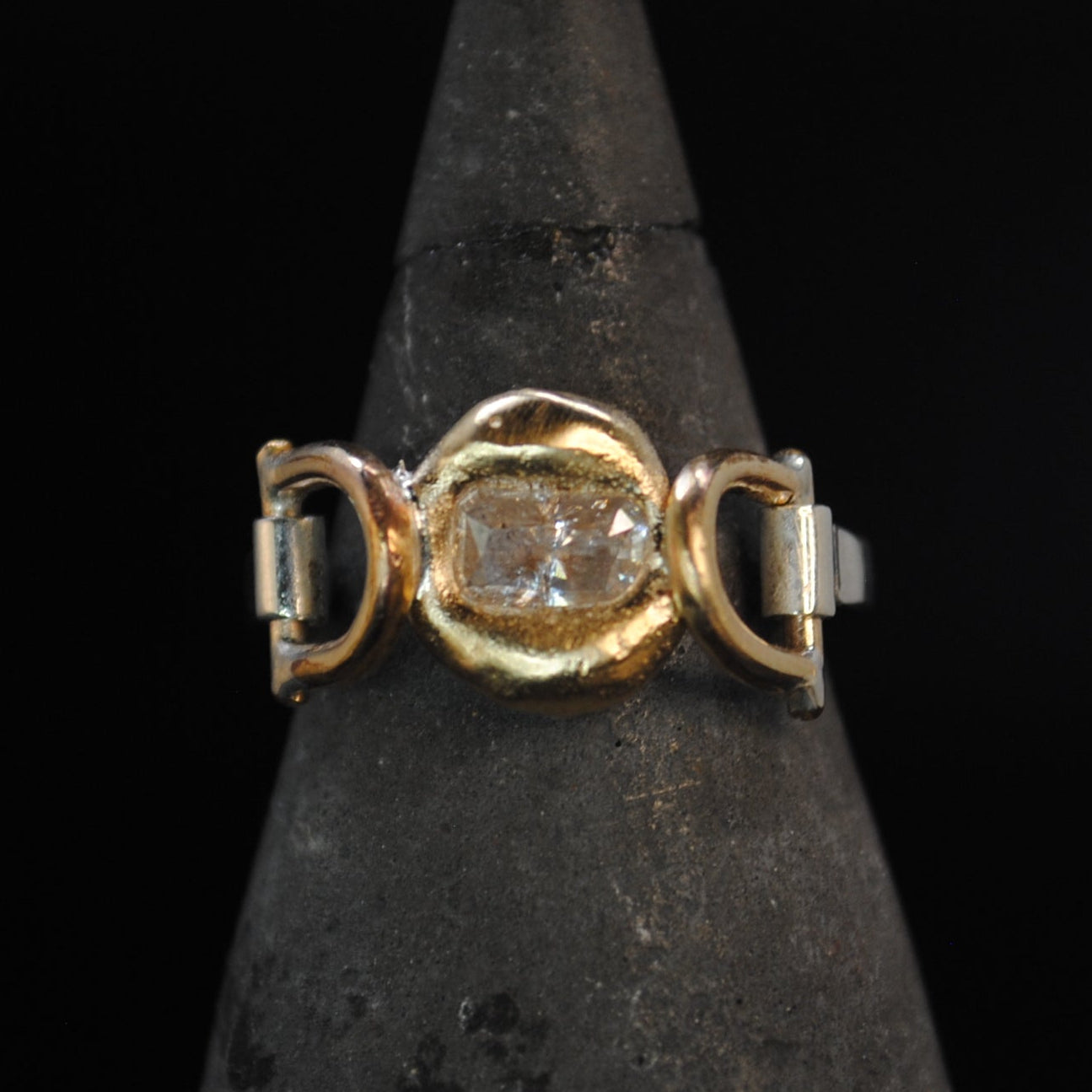 A gold ring with a central clear gemstone and side accents, displayed on a dark pointed jewelry cone against a black background.