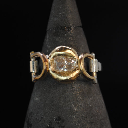 A gold ring with a central clear gemstone and side accents, displayed on a dark pointed jewelry cone against a black background.
