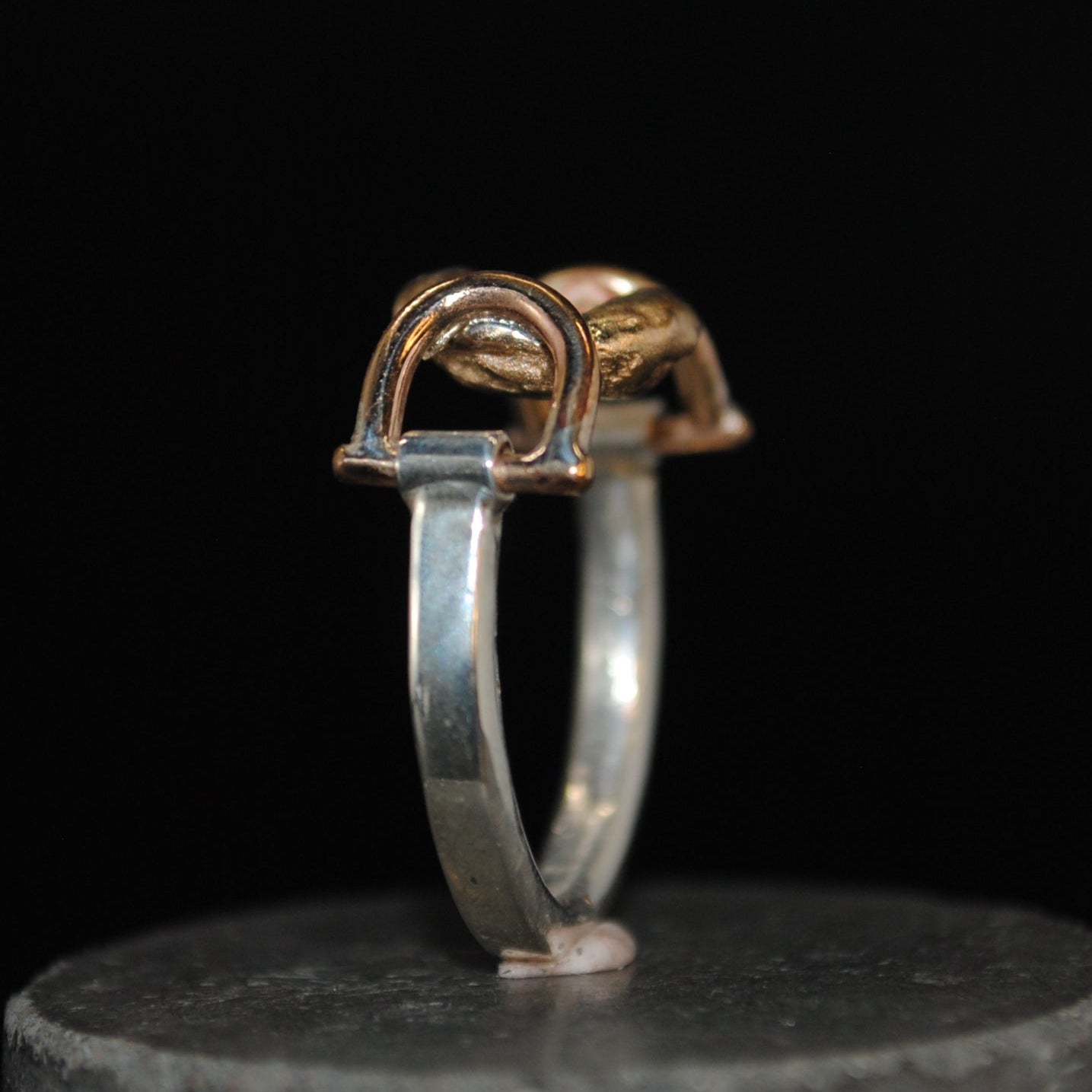 A two-tone ring with a silver band and a gold-toned horseshoe-shaped top, displayed on a dark gray cylindrical stand against a solid black background.