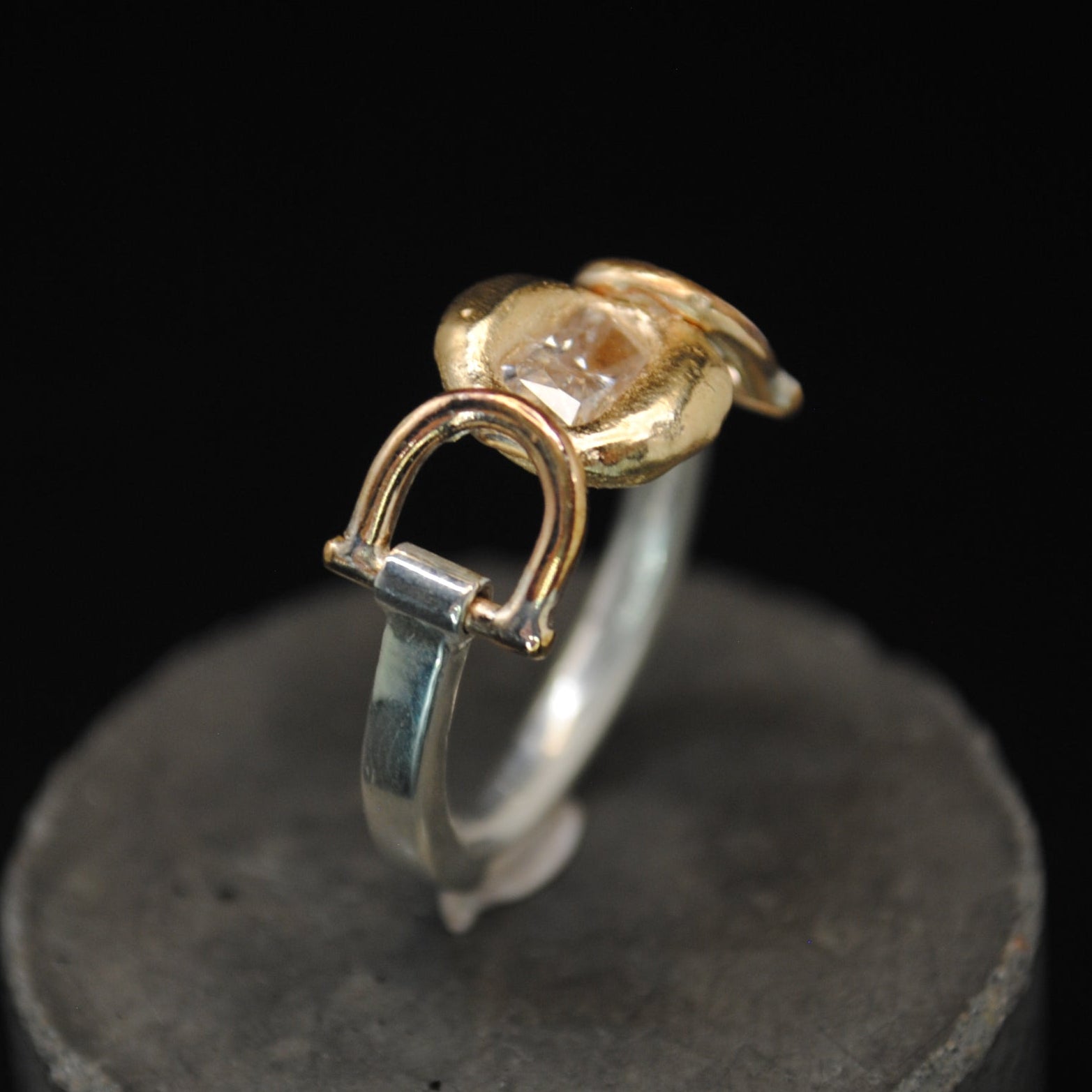 A two-tone ring with a gold-toned textured top and a silver-toned band, displayed on a dark gray cylindrical stand against a black background.