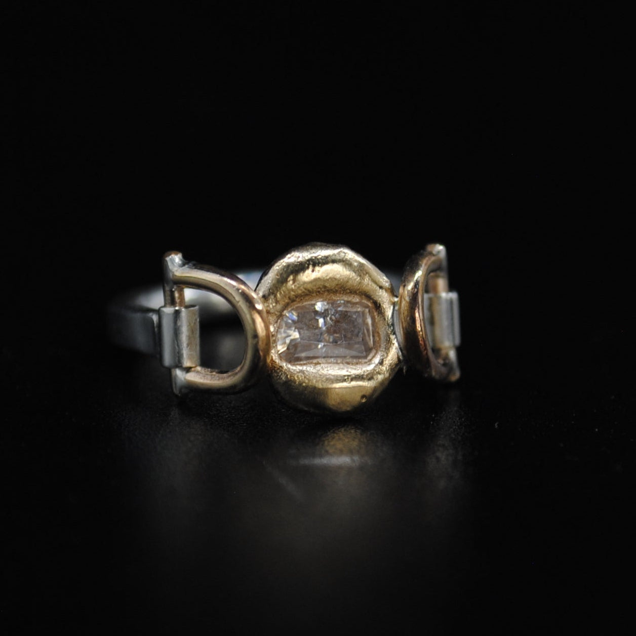 A gold and silver ring with a central diamond-cut gemstone, displayed on a reflective black surface.