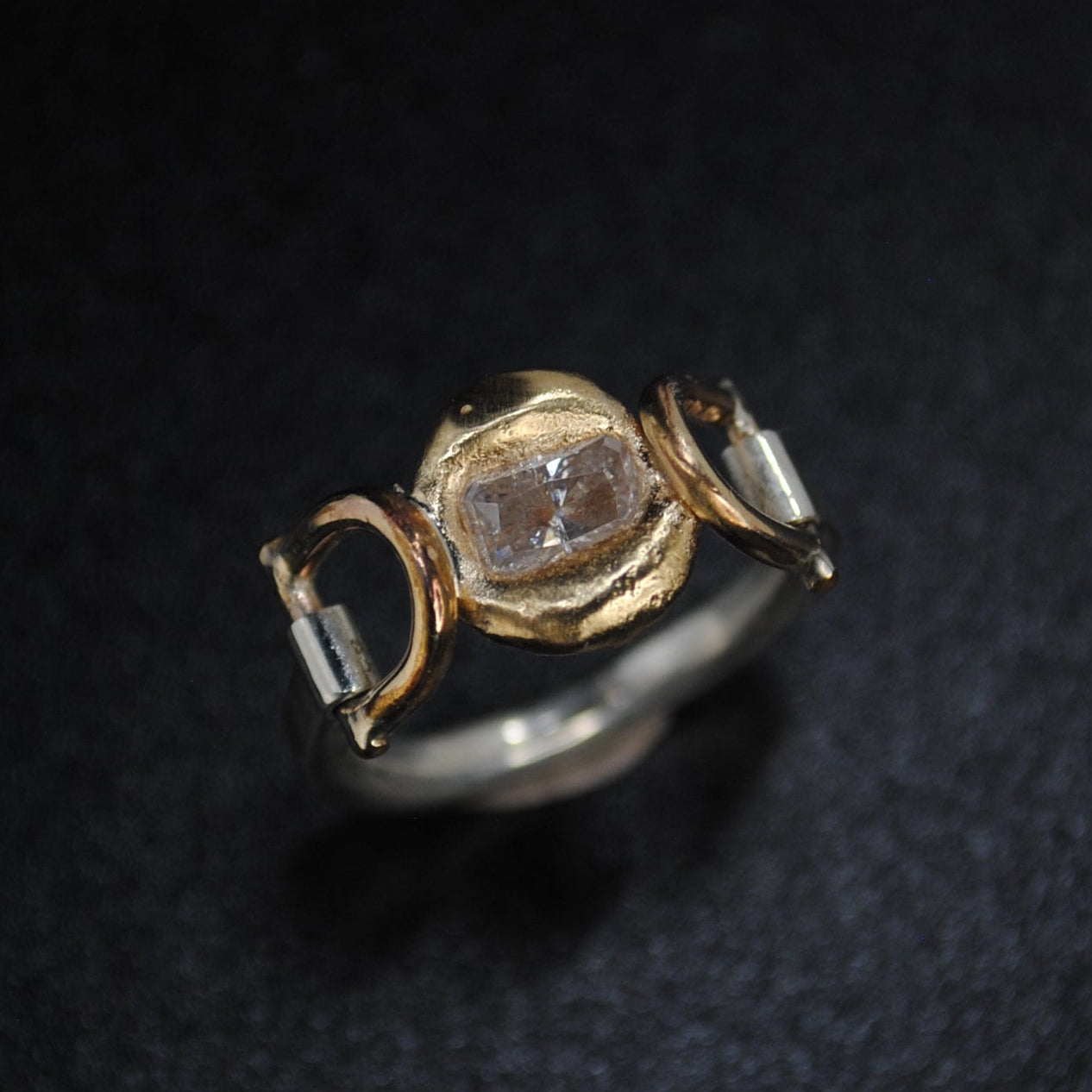 A vintage-style ring featuring a central oval clear gemstone in a gold-toned setting, flanked by two smaller clear stones in silver-toned settings, displayed on a dark textured background.