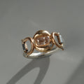 A diamond stone ring featuring a central rose gold cushion-cut gemstone flanked by hinges displayed on a gray surface with a soft shadow.