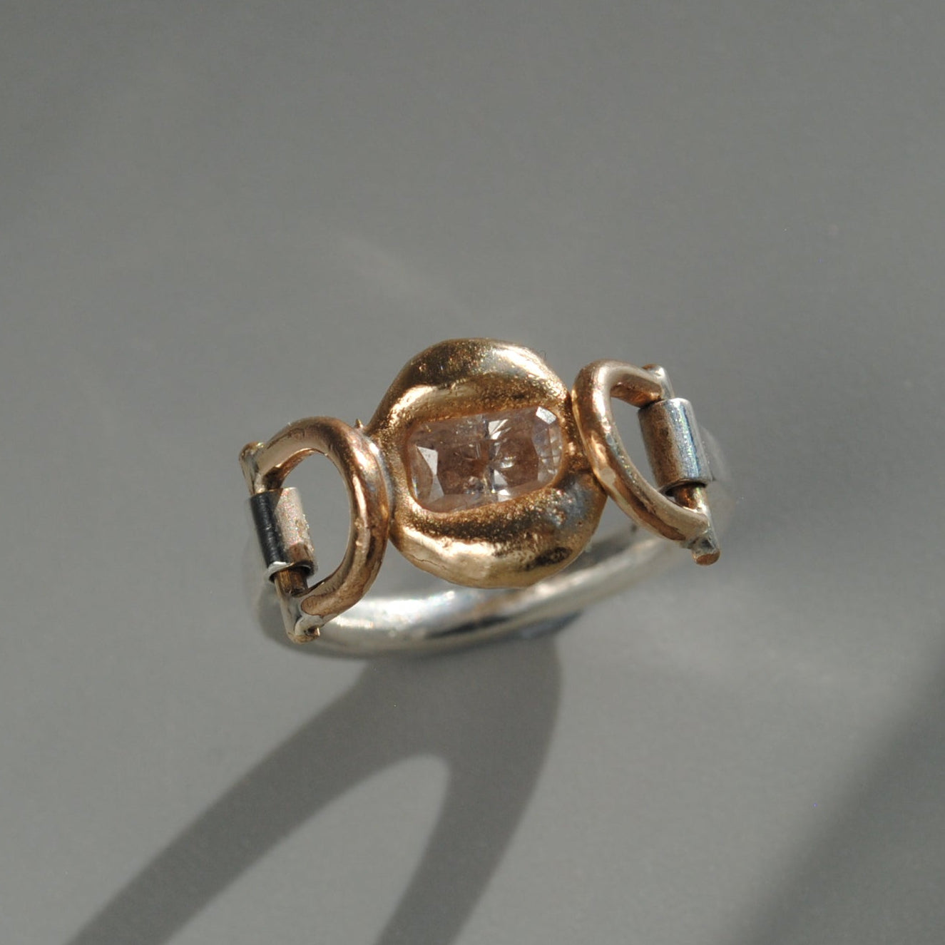 A diamond stone ring featuring a central rose gold cushion-cut gemstone flanked by hinges displayed on a gray surface with a soft shadow.
