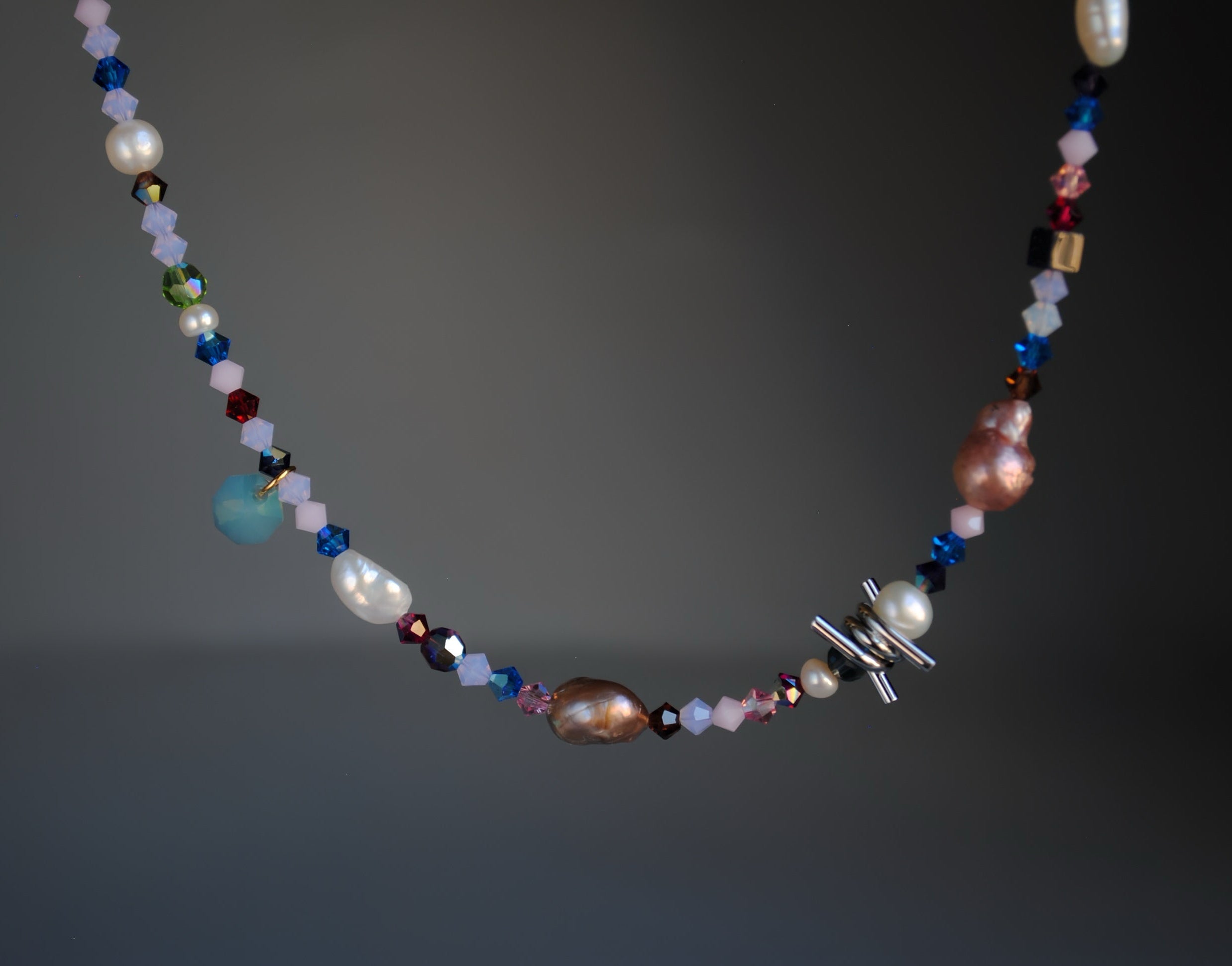 A beaded necklace featuring white pearls, faceted beads in blue, pink, purple, and brown, and a silver-toned cross charm, displayed against a dark gray gradient background.