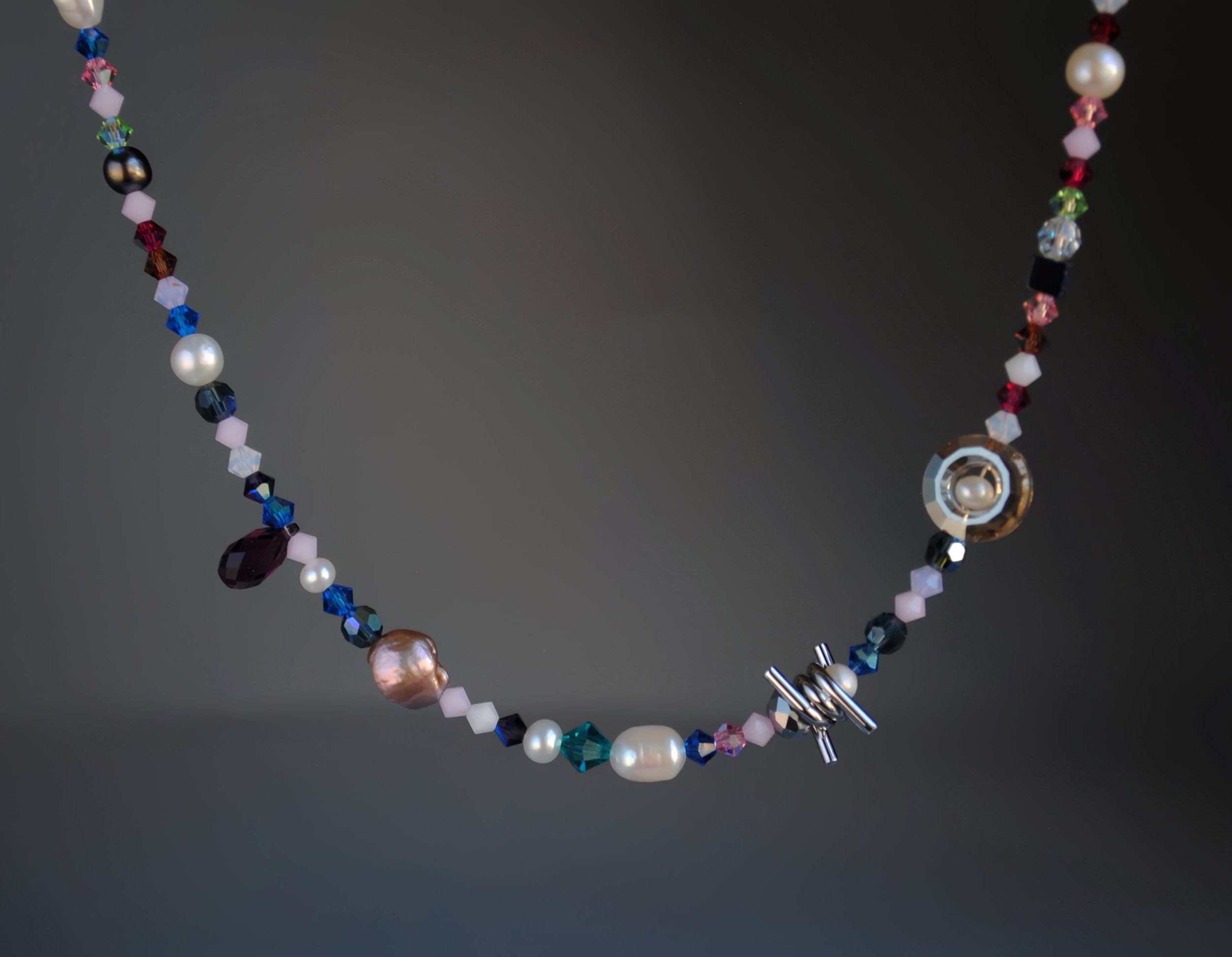 A beaded necklace featuring a mix of faceted crystal beads, faux pearls, and a silver-toned toggle clasp, displayed against a dark gray gradient background.
