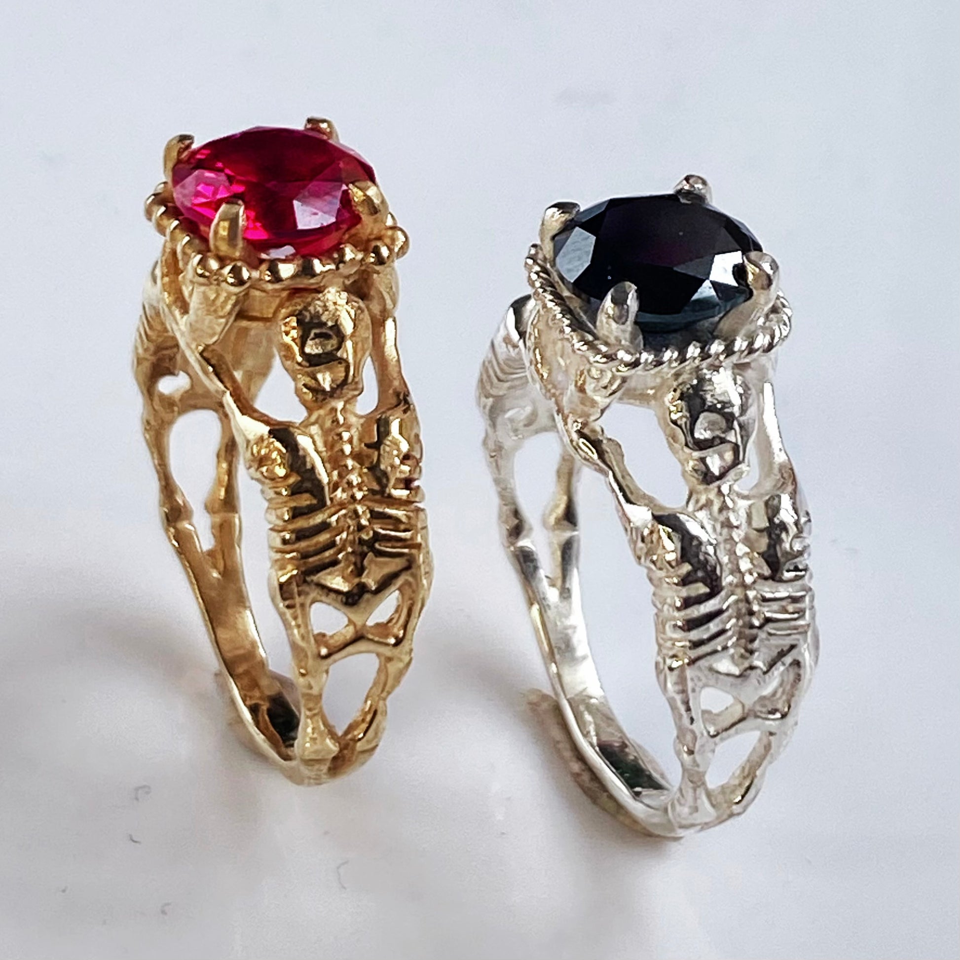 Two skeleton-themed rings, one gold with a red gem and one silver with a black gem, on a light gray background.