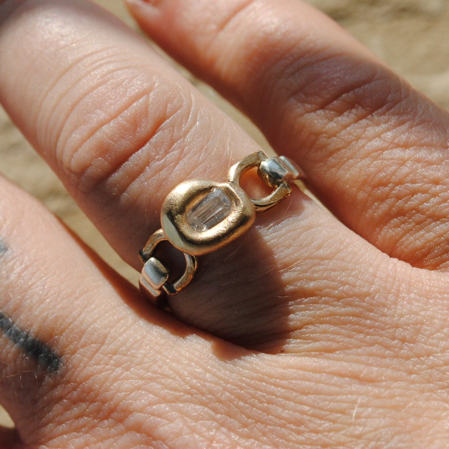 A gold ring with a unique interlocking double-oval design, worn on a finger with a cross tattoo, set against a blurred sandy background.
