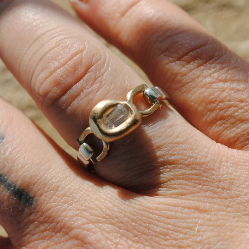 A gold ring with a unique interlocking double-oval design, worn on a finger with a cross tattoo, set against a blurred sandy background.