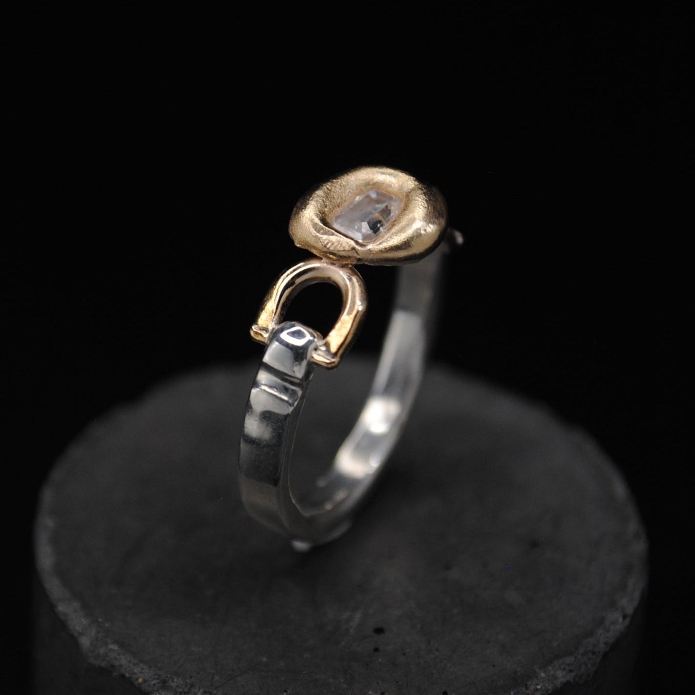 A two-tone ring with a silver band, a gold hanger clasp, and a small round gold bezel-set stone, displayed on a black cylindrical stand against a black background.