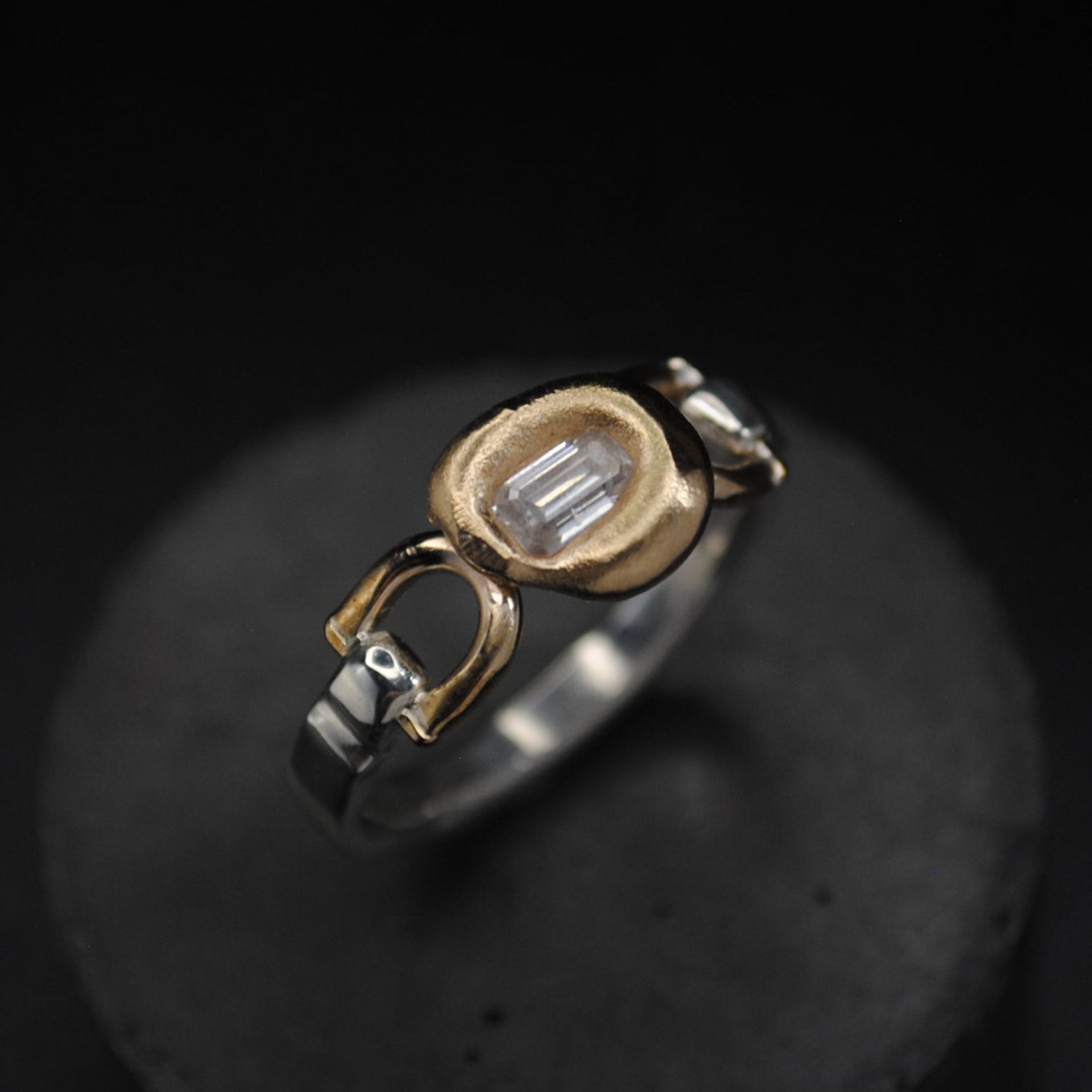 A two-tone ring with a gold oval bezel and a silver band, displayed on a round black stand against a dark background.