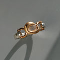 A two-tone ring featuring a textured rose gold central bezel with a clear baguette-cut gemstone, flanked by polished silver-colored metal sections, set against a neutral gray background.