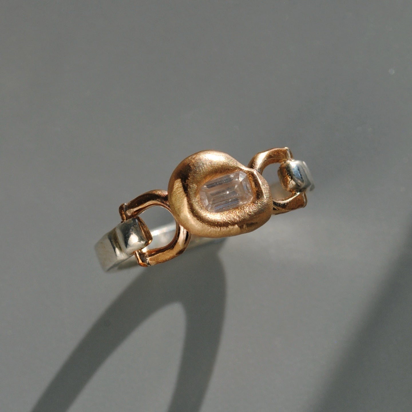 A two-tone ring featuring a textured rose gold central bezel with a clear baguette-cut gemstone, flanked by polished silver-colored metal sections, set against a neutral gray background.