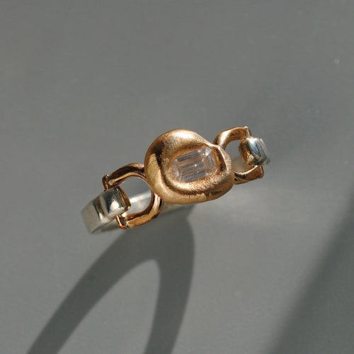 A two-tone ring featuring a textured rose gold central bezel with a clear baguette-cut gemstone, flanked by polished silver-colored metal sections, set against a neutral gray background.