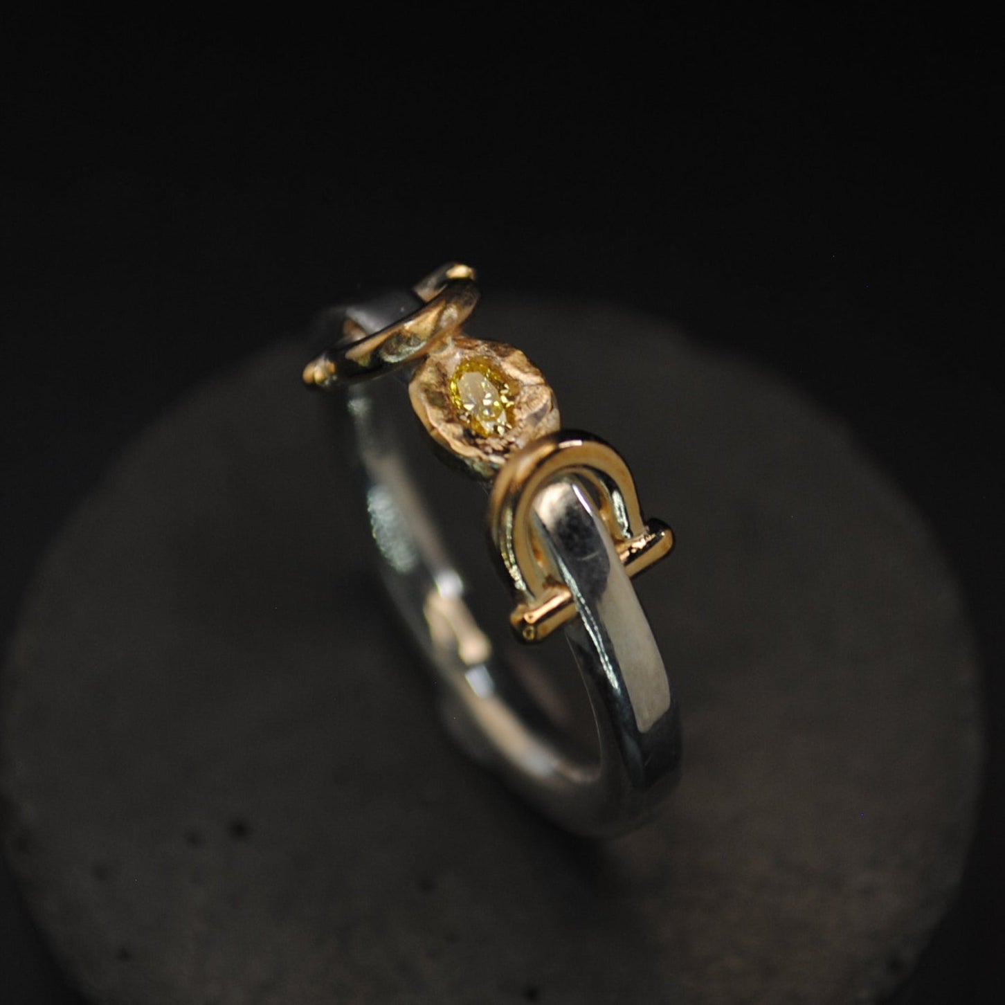 A two-tone metal ring with a gold and silver band, featuring a central gold element with a small clear gemstone, displayed on a dark textured stand against a black background.