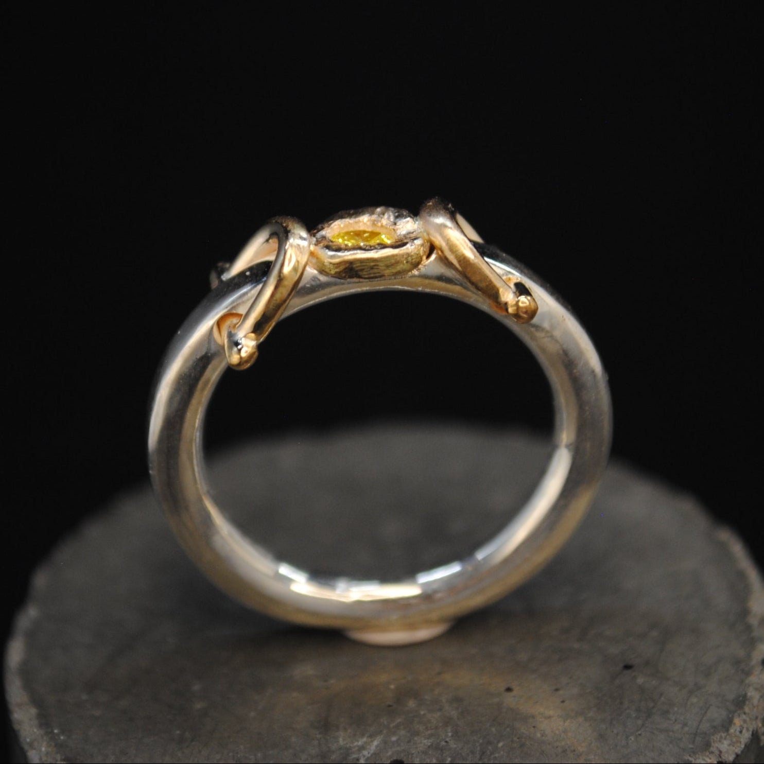 A silver ring with a gold-toned spider motif at the top, displayed on a dark textured surface against a solid black background.