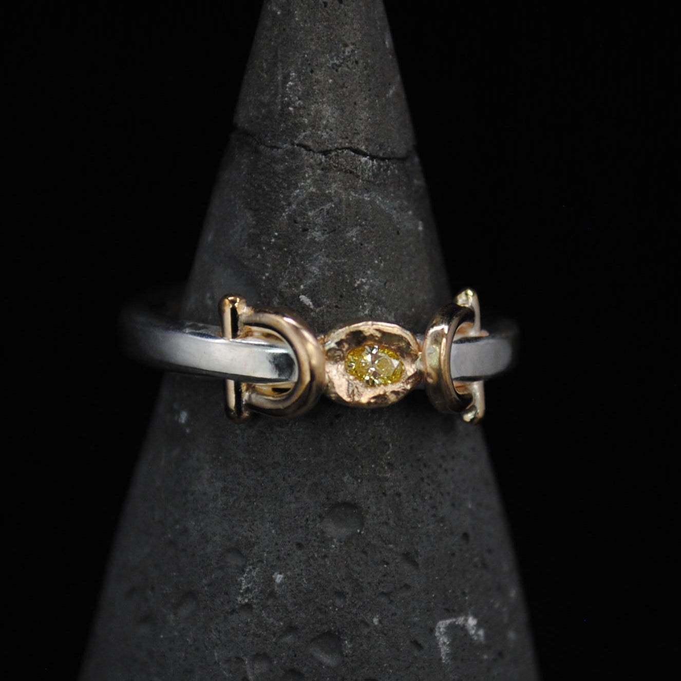 A delicate ring with a yellow gold center and a silver-toned band, displayed on a dark textured cone stand against a solid black background.