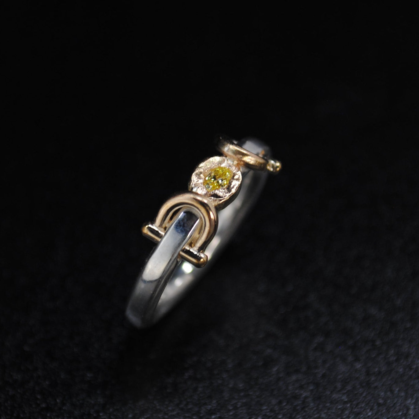 A two-tone ring with a silver band, a gold U-shaped accent, and a small round yellow gemstone, displayed on a dark textured background.