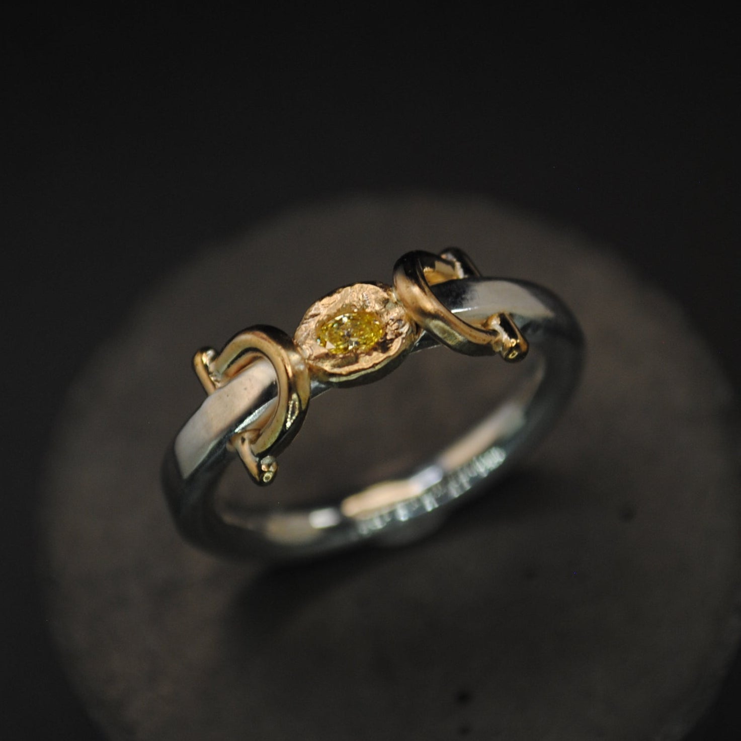 A unique ring featuring a central yellow diamond held in a gold setting, flanked by two curved silver bands with white enamel inlays, displayed on a dark textured stand against a black background.